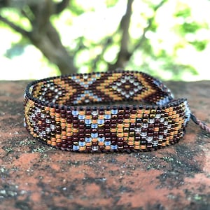 Op de afbeelding: Een bruin, gouden en witte kralen armband met een geometrisch patroon. De armband is gemaakt met kleine kralen en heeft een eenvoudig, elegant ontwerp.