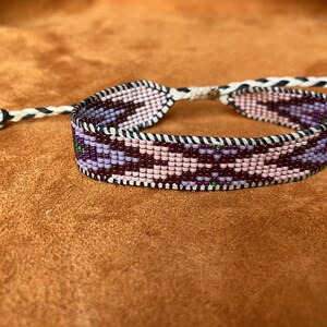 May include: A beaded bracelet featuring a pink, purple and brown geometric pattern. The bracelet is crafted with small beads and has a white cord tie.