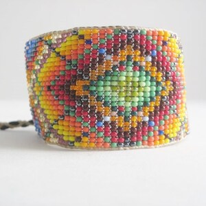 May include: A colorful beaded bracelet with a wide band design. The bracelet features a mosaic pattern of small beads in shades of red, orange, yellow, green, and blue. A woven cord is attached to the bracelet.