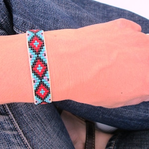 May include: A close-up of a beaded bracelet with a geometric pattern in red, blue, and black on a white background. The bracelet is worn on a person's wrist.