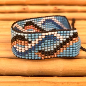 May include: A beaded bracelet with a geometric pattern in shades of blue, white, black, and copper. The bracelet is made with small, round beads and has a black cord closure.