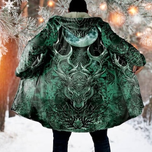 Pode incluir: Capa com capuz verde com um design detalhado de veado e uma lua crescente. A capa apresenta um forro de sherpa branco e padrões intrincados. O design é predominantemente verde e branco, com um fundo de floresta de inverno.