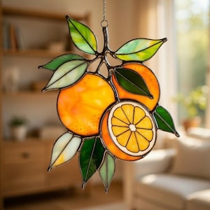 May include: Stained glass orange slice and oranges with green and yellow leaves. The decorative piece is suspended by a chain. The orange fruit is a bright orange color with a white rind and yellow center.