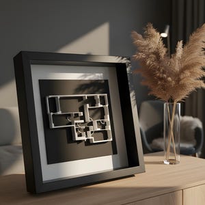 May include: A framed architectural floor plan in a black frame, featuring a white 3D model of a house layout against a black background. The frame is displayed on a wooden surface, with a vase of dried pampas grass in the background.