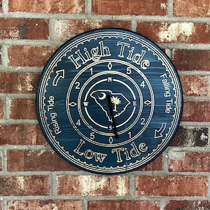 May include: A round, navy blue wooden tide clock with gold-colored engraved text and details. The clock face features the words "High Tide" and "Low Tide", along with a compass and the outline of South Carolina. The clock is mounted on a red brick wall.