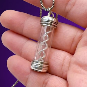 May include: A clear glass pendant necklace with a silver-colored DNA helix inside. The pendant has silver-colored metal caps on each end and hangs from a silver-colored chain. The background is a blurred purple.