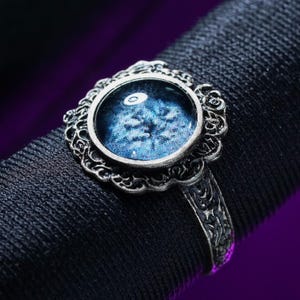 May include: A silver-toned ring with an ornate, filigree setting. The ring features a round, blue-toned stone with a detailed, eye-like pattern. The band has an engraved design, adding to the ring's intricate aesthetic.