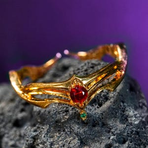 May include: A gold-colored tiara with a central teardrop-shaped red gemstone and a smaller green gemstone below. The tiara is set against a dark, textured background with a purple gradient.