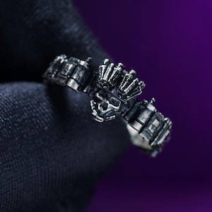 May include: A detailed silver ring featuring a skull design with intricate detailing. The ring has a dark, oxidized finish, and the skull is the focal point. The ring's design includes geometric shapes and a gothic aesthetic.