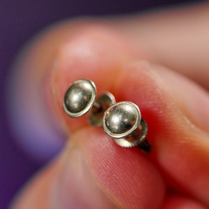 May include: A pair of small, silver-colored stud earrings. Each earring features a rounded, domed design. The earrings are held between fingers against a blurred background.