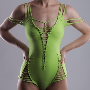 May include: A lime green bodysuit with a distinctive design. The one-piece garment has cut-out details on the sides and a central laced design. Thin straps extend over the shoulders and arms. The model is standing with hands on hips.