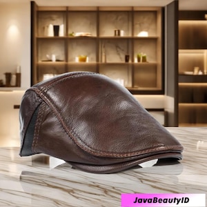 May include: A brown leather flat cap with a curved brim. The cap has a stitched design and a black accent on the side. The cap is sitting on a marble surface. The text "JavaBeautyID" is at the bottom.