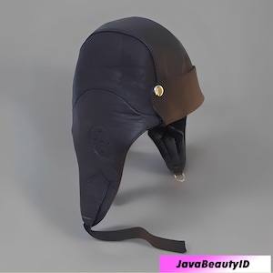 May include: A dark navy blue leather aviator hat with a brown visor. The hat has ear flaps and a chin strap. A gold-coloured button secures the visor to the hat. The hat is designed to protect the head and ears.