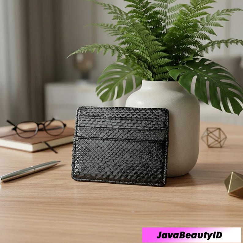 May include: A black cardholder with a snakeskin texture is displayed on a wooden table. The cardholder has several slots. A white vase with green plants, a pen, glasses, and a notebook are also on the table.
