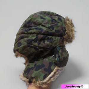 May include: A camouflage pattern trapper hat with faux fur trim and ear flaps. The hat features a green, brown, and black camouflage design, with two button closures on the ear flaps. The hat is designed to provide warmth and protection from the cold.