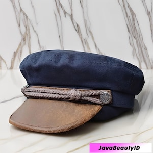 Puede incluir: Gorra de capitán azul marino con visera marrón. El sombrero presenta un detalle de cuerda trenzada y un botón plateado con un emblema de ancla. Un accesorio náutico clásico.