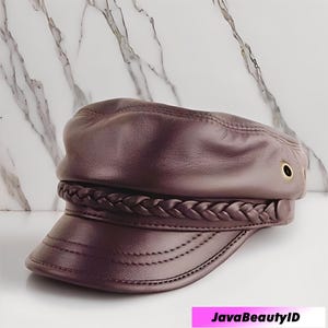 K&ouml;nnte beinhalten: Dunkelmalvenfarbene Lederkappe mit geflochtenem Band und kurzem Schirm. Die Kappe hat goldfarbene &Ouml;sen an der Seite. Der Hintergrund ist wei&szlig;er und grauer Marmor. Der Text "JavaBeautyID" befindet sich unten.