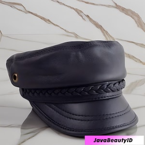 K&ouml;nnte beinhalten: Marineblaue Lederkappe mit geflochtenem Band und kleinem Schirm. Die Kappe hat ein gen&auml;htes Design und eine kleine goldfarbene &Ouml;se an der Seite. Der Text "JavaBeautyID" ist unten im Bild sichtbar.