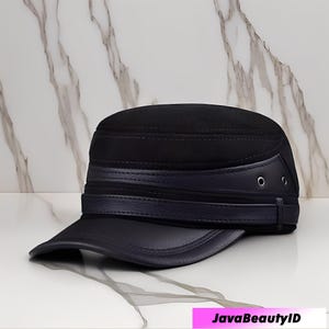 May include: A black military-style cap with a flat brim. The cap features a dark blue leather band and brim, with two small metal eyelets on the side. The top of the cap has a textured, suede-like appearance.