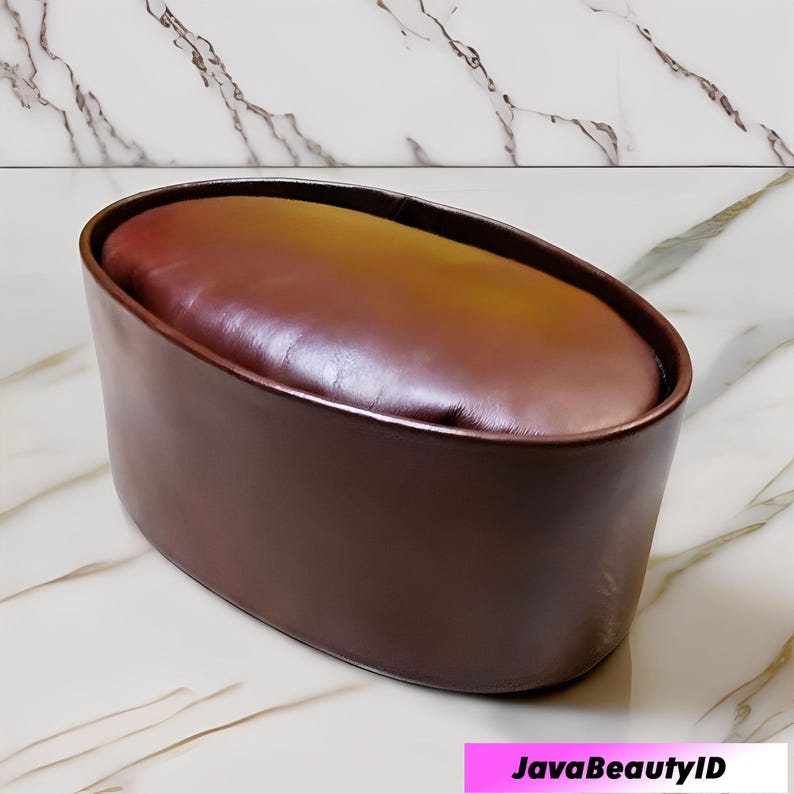 Peut inclure: Un pouf ovale marron avec un dessus en cuir et un ext&eacute;rieur marron fonc&eacute; assorti. Le pouf est pos&eacute; sur un fond &agrave; motifs de marbre. Le texte "JavaBeautyID" est visible dans le coin inf&eacute;rieur droit.