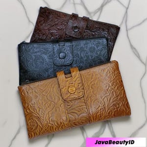 Pode incluir: Três carteiras de couro em tons de castanho, preto e bege, cada uma com um padrão floral em relevo. As carteiras têm formato retangular com fecho de aba e botão. As carteiras estão dispostas sobre uma superfície de mármore branco.