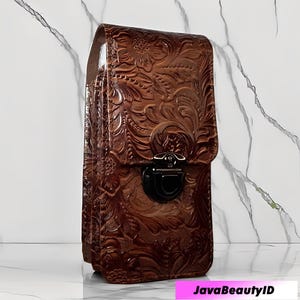 Può includere: Una custodia per telefono in pelle marrone con un design floreale in rilievo. La custodia ha una chiusura a patta con chiusura nera e un cinturino. La custodia è realizzata in pelle marrone scuro e ha una finitura lucida.