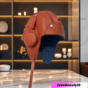 Può includere: Un cappello da aviatore in pelle marrone con paraorecchie e sottogola. Il cappello ha una fodera interna blu scuro e rivetti color argento. Il cappello è progettato per coprire le orecchie e il collo, offrendo protezione dagli elementi. Stile vintage.