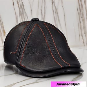 May include: A black leather newsboy cap with red stitching. The cap has a rounded crown and a small, stiff brim. The stitching creates a decorative pattern on the cap's surface. The cap is displayed on a marble surface.