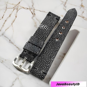 Może przedstawiać: Czarny pasek do zegarka z teksturowanym, kropkowanym wzorem. Pasek ma srebrną klamrę i trzy małe, okrągłe nity. Pasek leży na białej powierzchni z marmurowym wzorem.