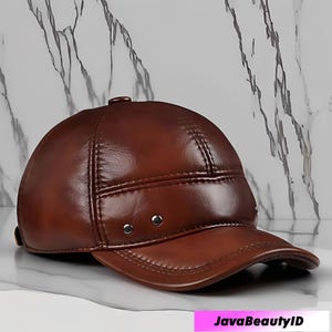 May include: A brown leather cap with a curved brim. The cap features a rounded crown with stitched details and small silver rivets. The cap is made of a shiny, smooth leather material.