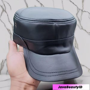 May include: A black leather military-style cap with a flat top and a short, curved brim. The cap features a band around the crown and is held by a hand. The text "JavaBeautyID" is at the bottom.