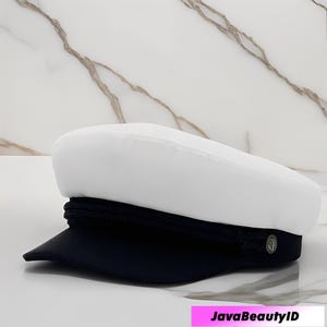 Peut inclure: Casquette de marin blanche et bleu marine, avec une couronne arrondie et une visière courte et rigide. Le chapeau présente une bande tressée et un petit emblème décoratif. Fond en marbre blanc.