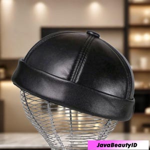 May include: A black, leather-look cap with a rounded crown and a short, folded brim. The cap has a small button detail on top and is displayed on a wire form. The cap appears to be new.