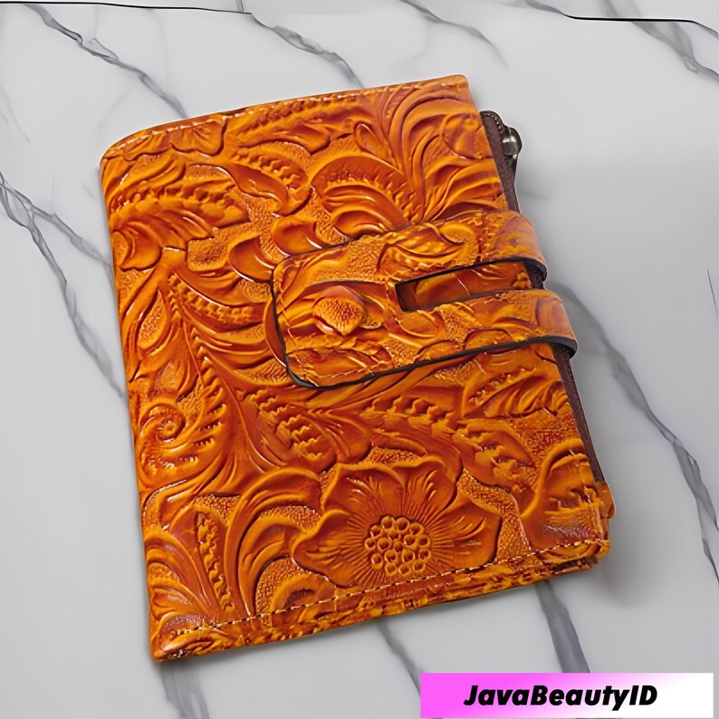 May include: An orange leather bi-fold wallet featuring a floral embossed pattern. The wallet has a snap closure and a zip. The floral design covers the entire surface. The background is a white marble surface.