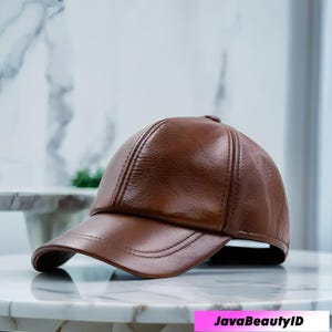 Puede incluir: Gorra de béisbol de cuero marrón con visera curva. La gorra tiene un acabado liso y brillante y detalles de costura visibles. El texto "JavaBeautyID" es visible en la esquina inferior derecha.
