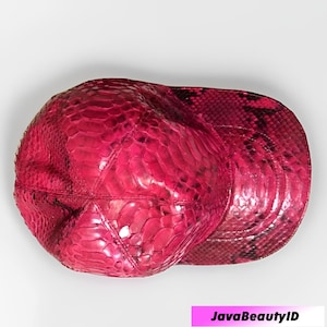 Può includere: Un berretto da baseball rosa acceso con un motivo a pelle di serpente. Il berretto presenta una visiera curva e una corona strutturata, che mostra una texture lussuosa e un colore audace. Un accessorio di moda.