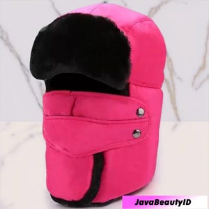 May include: A vibrant pink winter hat with a black faux fur trim. The hat features a quilted design and ear flaps that can be secured with two silver-colored buttons. The text "JavaBeautyID" is visible in the bottom right corner.