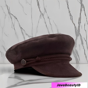 Puede incluir: Gorra de pana marrón con visera corta. La gorra presenta un detalle decorativo de cuerda y un botón en el lateral. Un sombrero de estilo clásico, adecuado para varios atuendos.