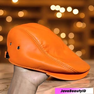 以下が含まれることがあります： 鮮やかなオレンジ色のニュースボーイキャップ。 つばがカーブしており、滑らかな素材で作られています。 小さな金属製の留め金が付いています。 無地でクラシックなデザインの帽子です。