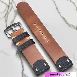 May include: Two-piece watch strap in brown leather with a black textured accent. The strap features a silver-colored buckle and is designed with multiple adjustment holes. The watch strap is displayed on a marble surface.