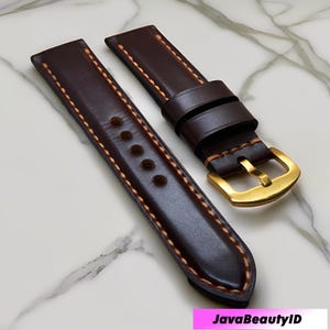 May include: Two dark brown leather watch straps with tan stitching. One strap has a gold-coloured buckle. The straps are displayed on a white and grey marble surface. The text "JavaBeautyID" is visible.