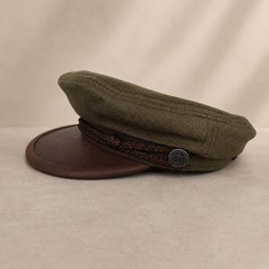 May include: Olive green cap with a brown leather brim and a decorative brown rope detail. The cap features a silver button on the side. The hat is a classic style, suitable for various occasions.