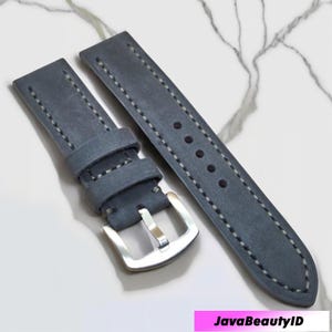 May include: Two-piece watch strap in a muted blue-gray color. The straps are made of a textured material with white stitching along the edges. A silver-colored buckle is attached to one strap. The text "JavaBeautyID" is in the bottom right corner.