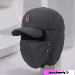 May include: A dark gray winter hat with a quilted pattern, ear flaps, and a face covering. The hat has a small brim and a rectangular patch with text. The hat is designed for cold weather.