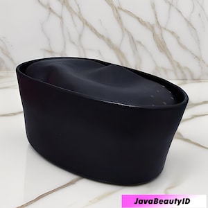 Peut inclure: Un r&eacute;cipient noir de forme ovale pour produits de beaut&eacute;, avec un dessus incurv&eacute;. Le r&eacute;cipient a une finition lisse et mate et repose sur une surface en marbre blanc et gris. Le texte "JavaBeautyID" est affich&eacute; dans un rectangle rose en bas.