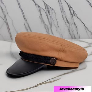 May include: Tan-colored captain's hat with a black visor and a black rope detail. The hat features a decorative button on the side. The hat is a stylish accessory.