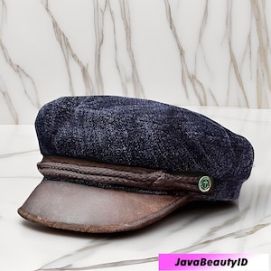 Może przedstawiać: Granatowa czapka marynarska z brązowym skórzanym daszkiem i plecionym wykończeniem. Czapka posiada mały zielony guzik z emblematem kotwicy. Stylowy dodatek.