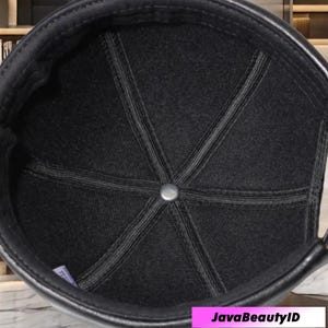 May include: Black cap with a leather brim and a textured fabric crown. The crown is divided into panels with visible stitching, meeting at a central metal button. The interior is lined with a dark fabric.