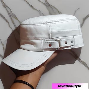 May include: A white leather military-style cap with a short brim. The cap features a decorative strap with a silver buckle and rivets. The cap is held in a hand.