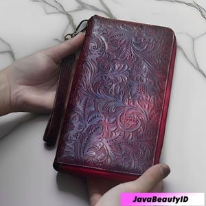Pode incluir: Uma carteira de couro bordô com um design floral em relevo. A carteira tem fecho de zíper e uma alça de pulso. O padrão floral é detalhado e cobre toda a superfície. A carteira é segurada em duas mãos.